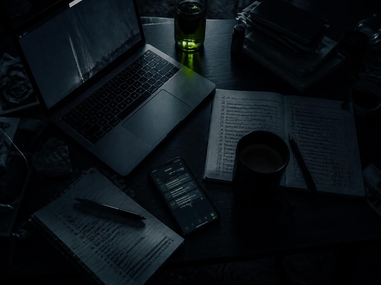 Mesa de trabalho à noite com laptop, celular e listas inacabadas